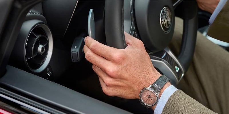 A driver's hand using silver paddle shifters on an Alfa Romeo steering wheel.